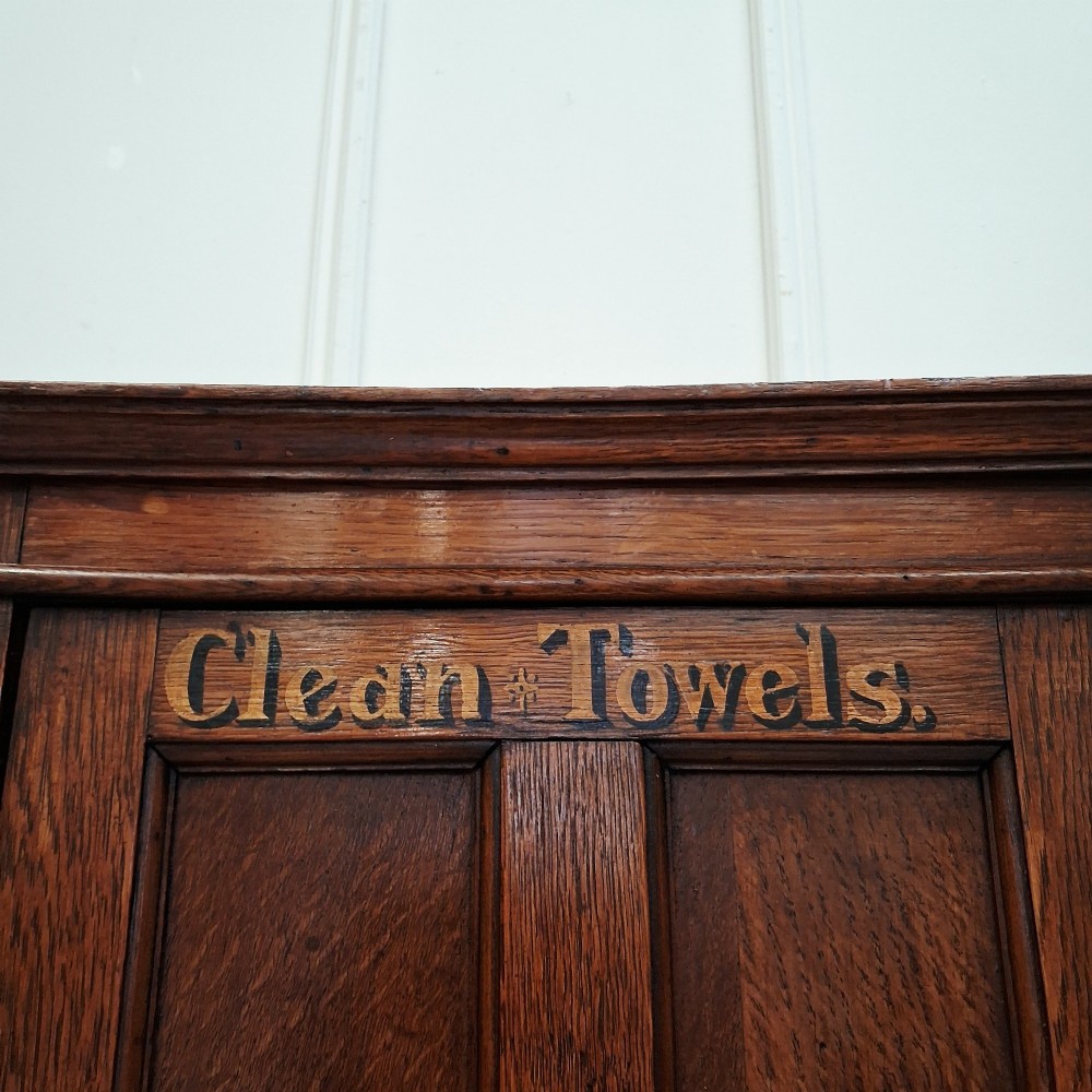 An antique medium brown oak wall cupboard featuring a panelled single door with Clean Towels in gilt lettering across the top and a brass door knob. View II