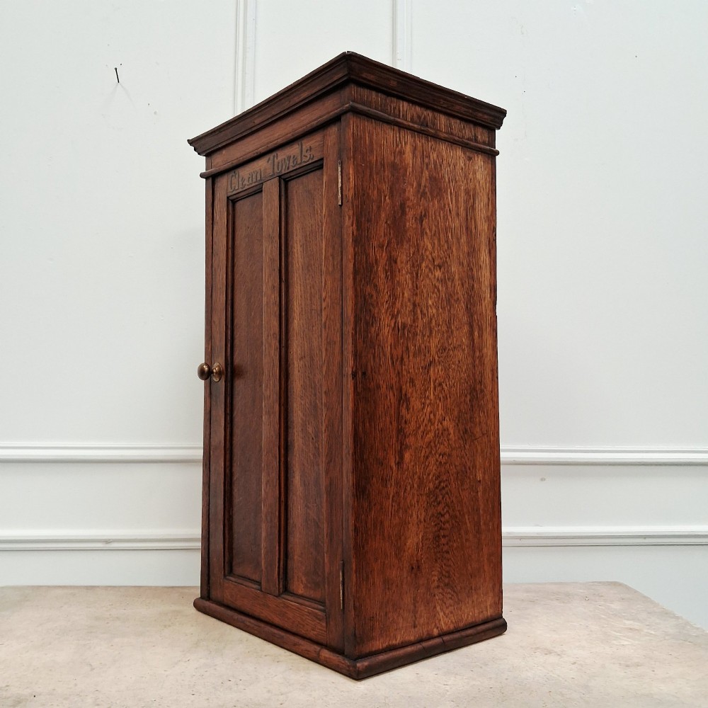 An antique medium brown oak wall cupboard featuring a panelled single door with Clean Towels in gilt lettering across the top and a brass door knob. View IV