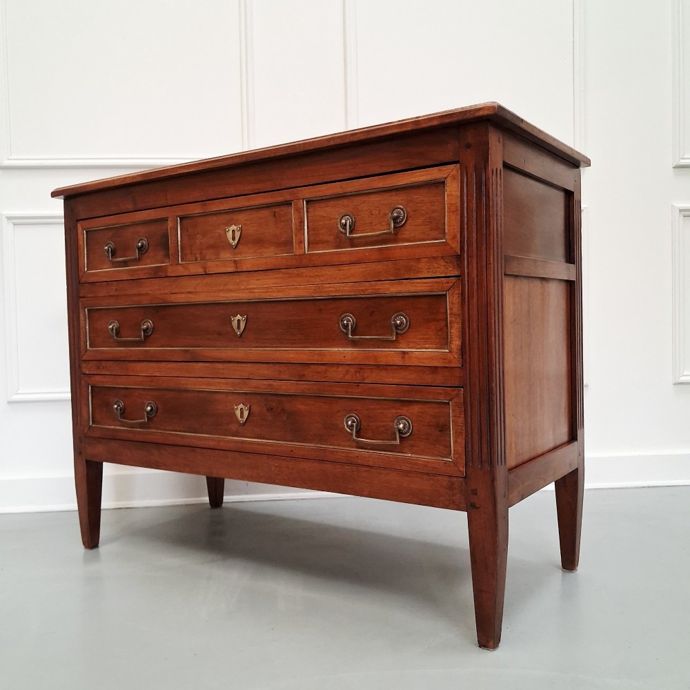 A late 19th-century French walnut commode featuring three drawer tiers, brass handles, shield-shaped escutcheons, and tapered fluted legs. View II