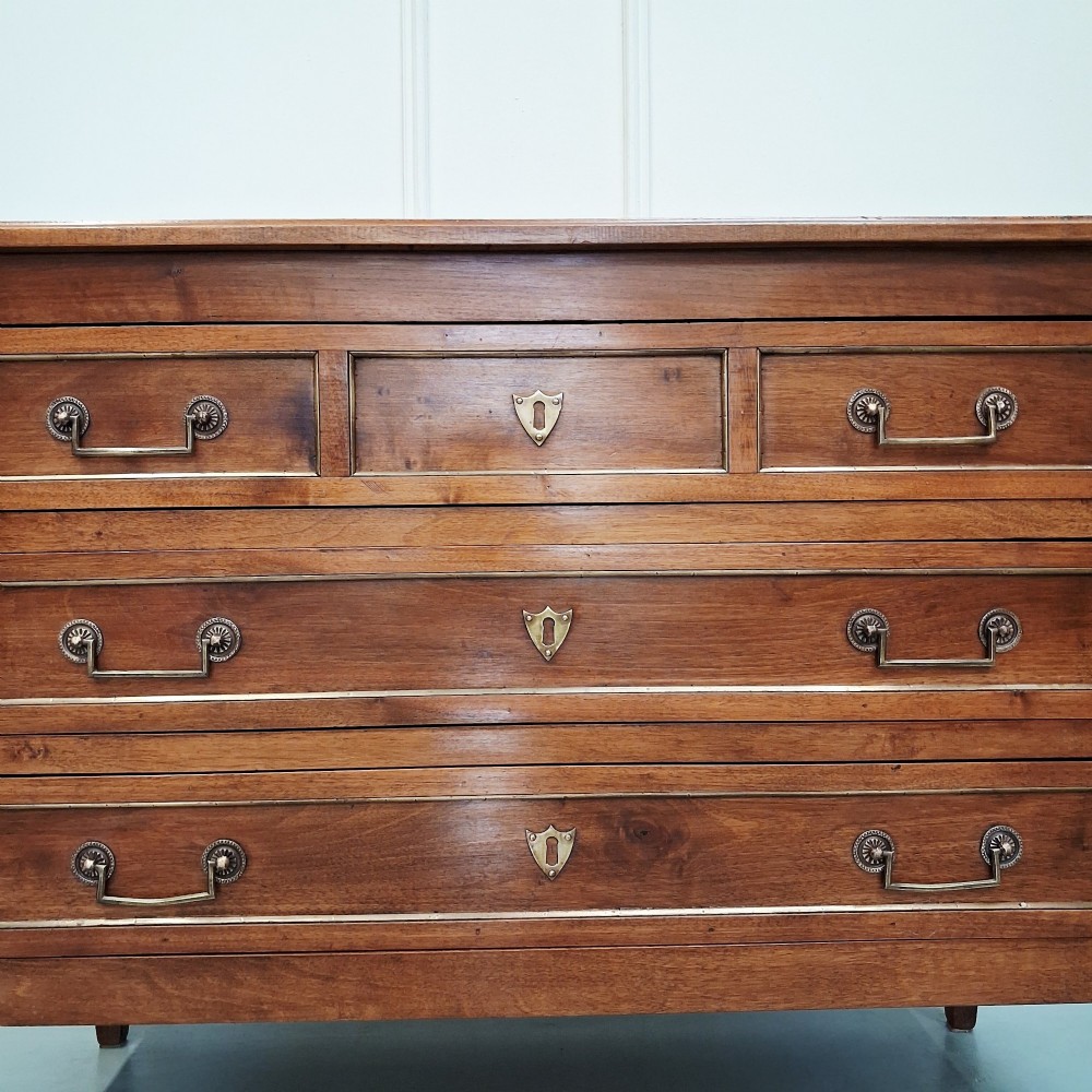 A late 19th-century French walnut commode featuring three drawer tiers, brass handles, shield-shaped escutcheons, and tapered fluted legs. View IV