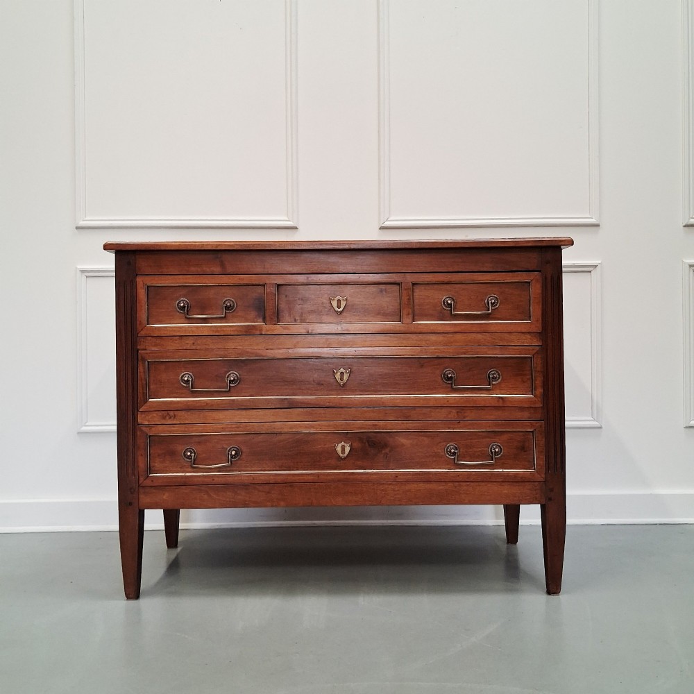 A late 19th-century French walnut commode featuring three drawer tiers, brass handles, shield-shaped escutcheons, and tapered fluted legs. View V