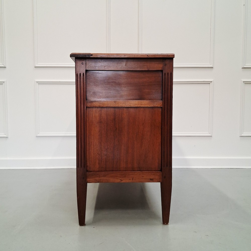 A late 19th-century French walnut commode featuring three drawer tiers, brass handles, shield-shaped escutcheons, and tapered fluted legs. View VI