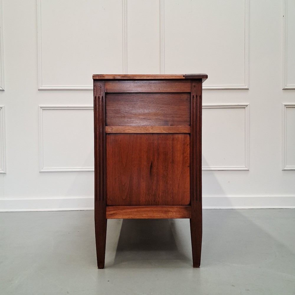 A late 19th-century French walnut commode featuring three drawer tiers, brass handles, shield-shaped escutcheons, and tapered fluted legs. View VIII