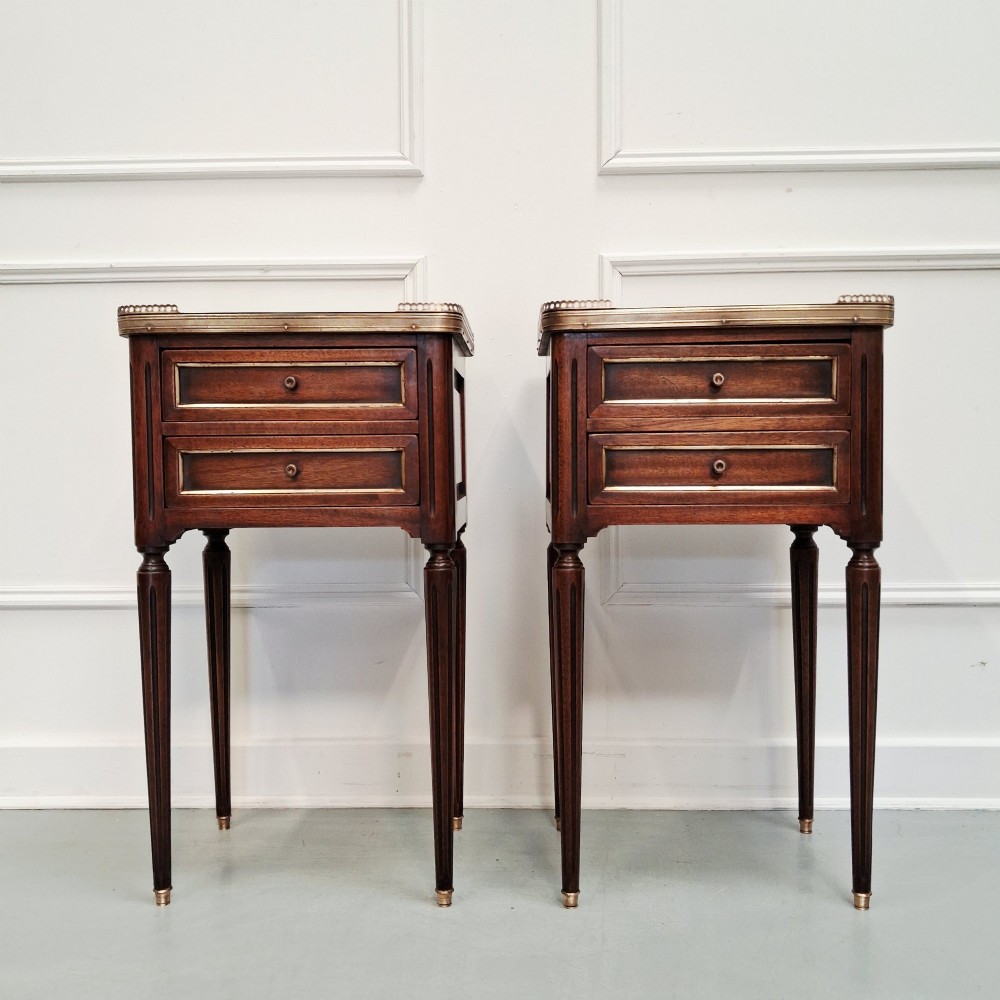 A pair of slim dark wood French bedside tables featuring fluted legs, brass galleries, two drawers, and inset marble tops.