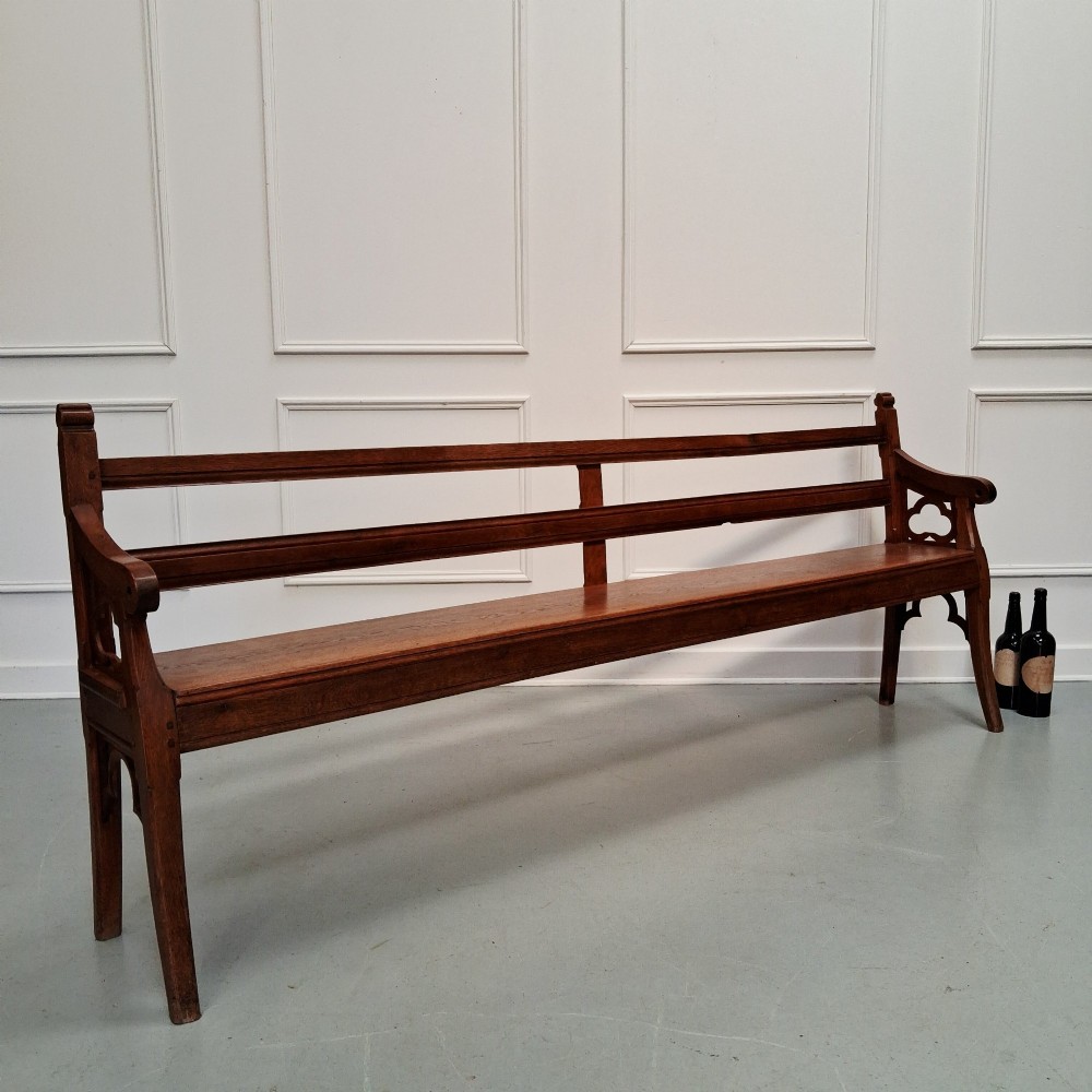 A long antique dark oak bench featuring an open rail backrest, sculpted armrests with trefoil cutouts, and subtle Gothic arch details beneath the frame. View IV