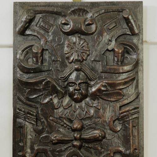 fig 1. A rectangular dark oak timber panel featuring a high-relief hand carving of a winged cherub head surrounded by intricate scrollwork and geometric strapwork.