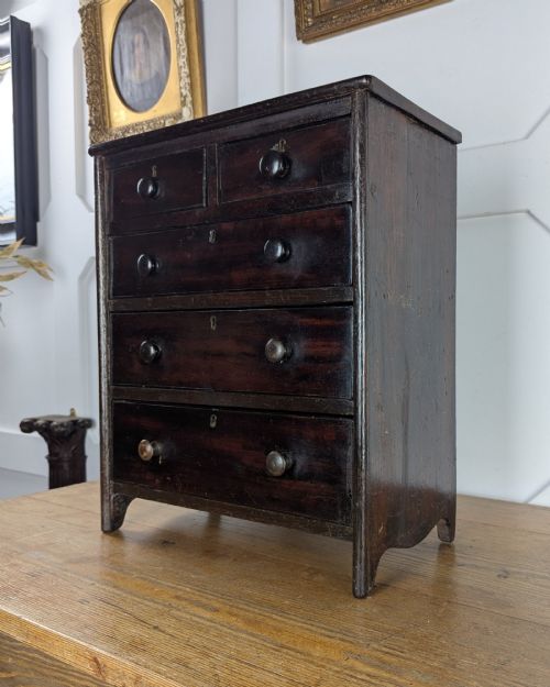fig 1. A 19th-century miniature wooden chest of drawers with a dark stained finish, two small top drawers over three long drawers, turned knobs, and brass escutcheons.