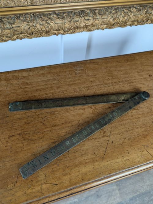 fig 1. A solid brass folding ruler featuring stamped block text reading WINDSOR BURIAL BOARD resting on a wooden surface.