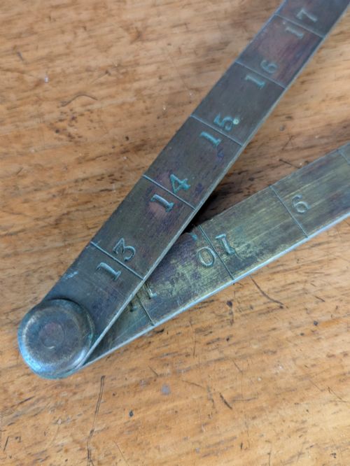 fig 2. A solid brass folding ruler featuring stamped block text reading WINDSOR BURIAL BOARD resting on a wooden surface.