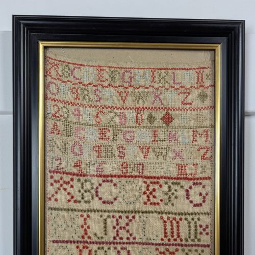 fig 1. A vertical framed Georgian needlework sampler featuring rows of cross-stitched alphabets, numbers, and urn and bird motifs on a natural linen fabric, housed in a dark frame with a gilt inner slip.