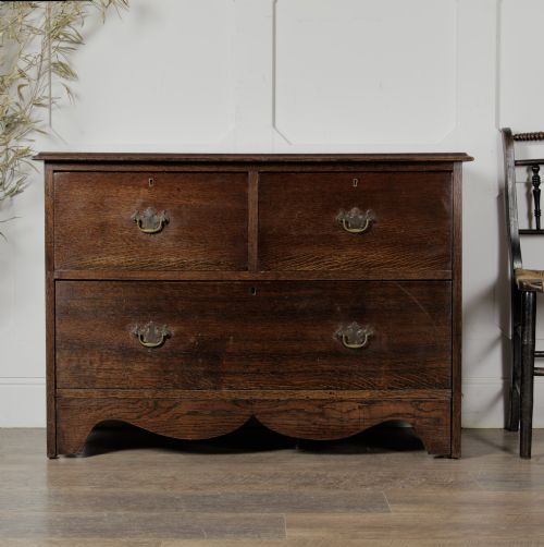 fig 1. A dark solid oak chest of drawers featuring two upper drawers over a wide lower drawer with brass swan-neck handles and a shaped bottom apron.