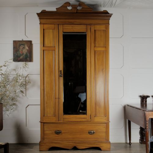 fig 2. An early 20th-century satinwood wardrobe featuring a central mirrored door, carved top pediment, panelled sides, and a single lower drawer with metal drop handles.