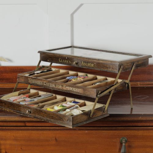 fig 1. A dark wooden multi-tiered cantilever haberdashery display box with a glass lid bearing a yellow anchor logo, shown open with thread spools inside.