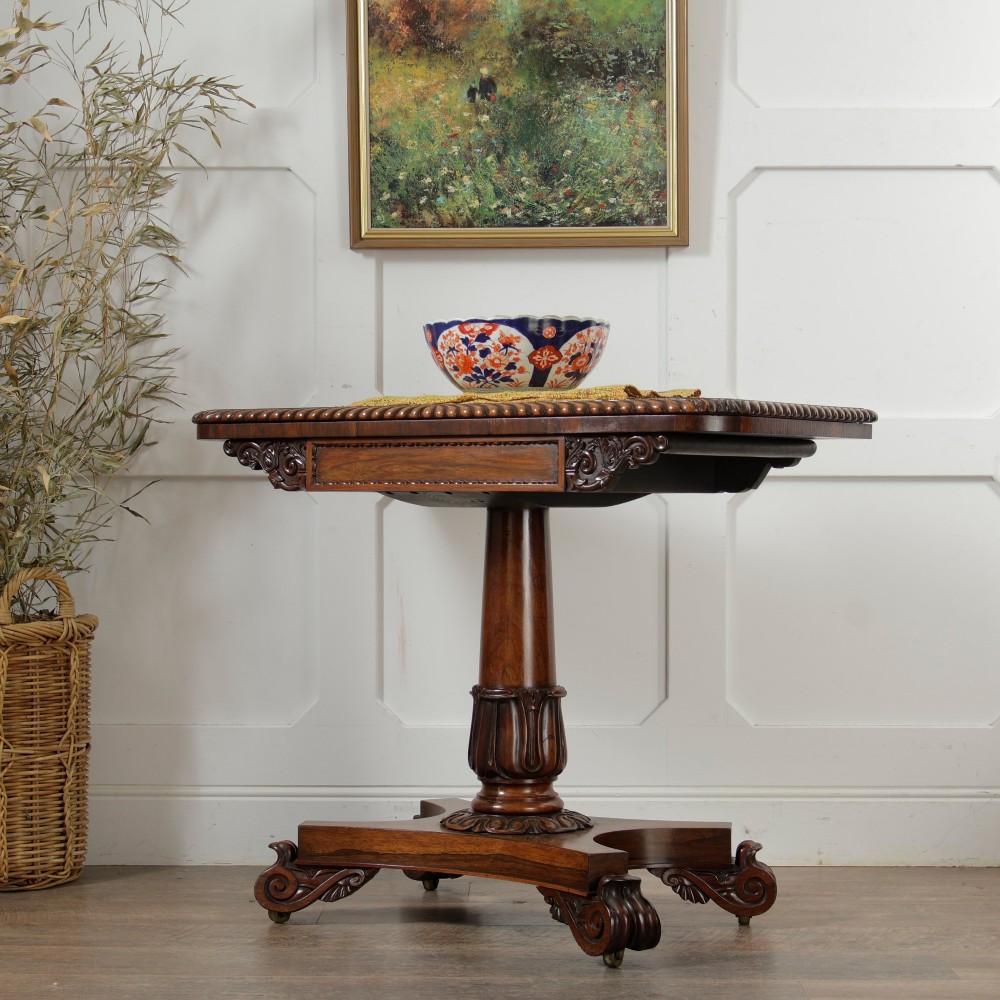 A William IV dark rosewood card table featuring a pedestal stem, carved scroll feet on castors, and a gadrooned top edge.