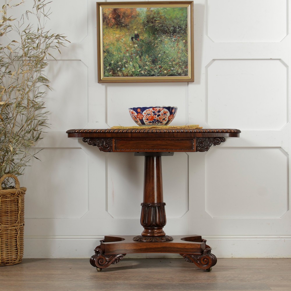 A William IV dark rosewood card table featuring a pedestal stem, carved scroll feet on castors, and a gadrooned top edge. View II