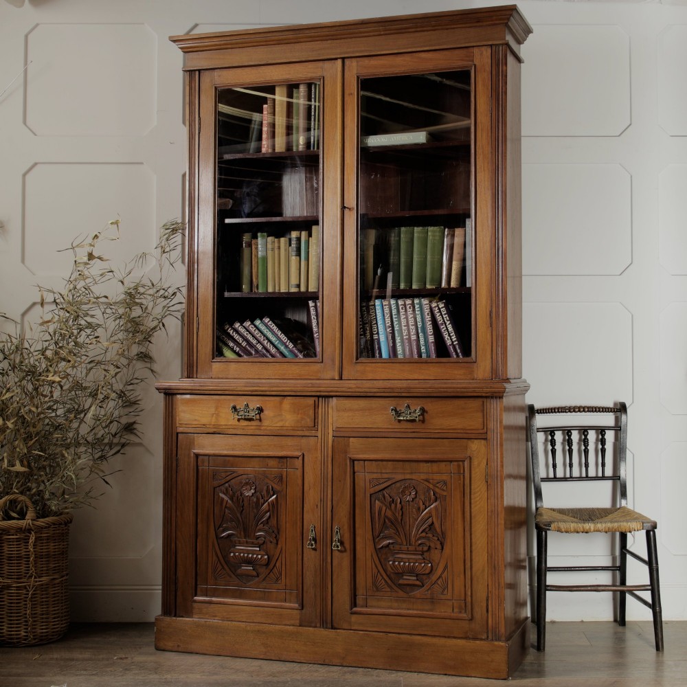 A late Victorian wooden glazed bookcase featuring two upper glass doors above two drawers and a double-door cupboard carved with floral urn motifs.