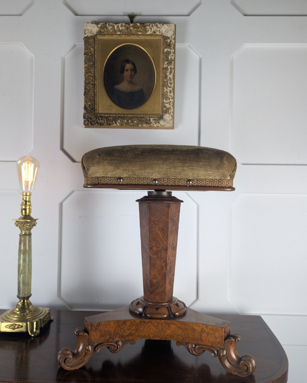 An antique William IV figured walnut revolving piano stool featuring an olive green upholstered square seat, a faceted column, and a carved triform base with scroll feet. View II
