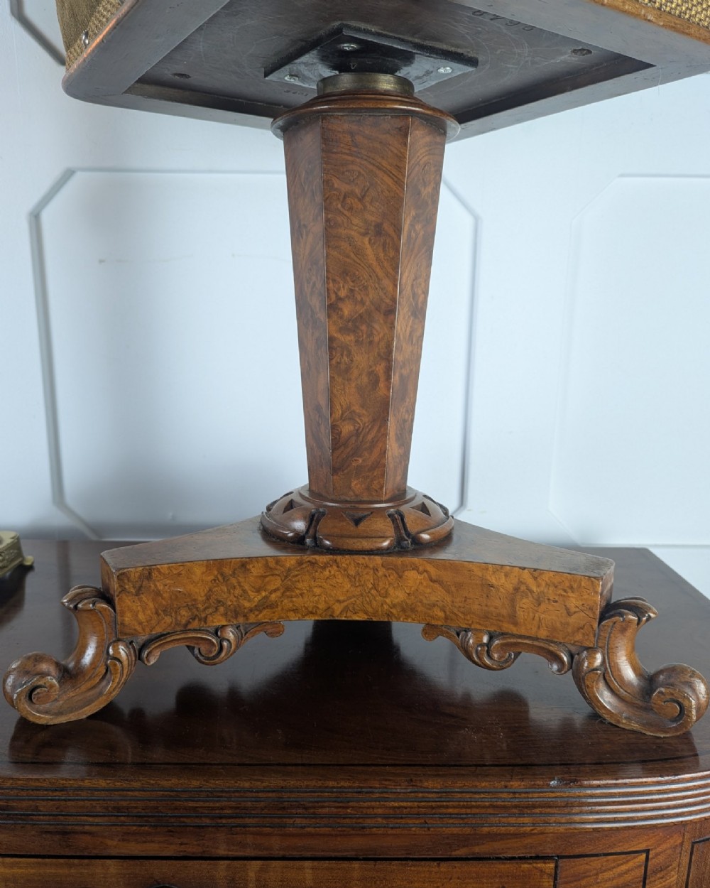 An antique William IV figured walnut revolving piano stool featuring an olive green upholstered square seat, a faceted column, and a carved triform base with scroll feet. View V