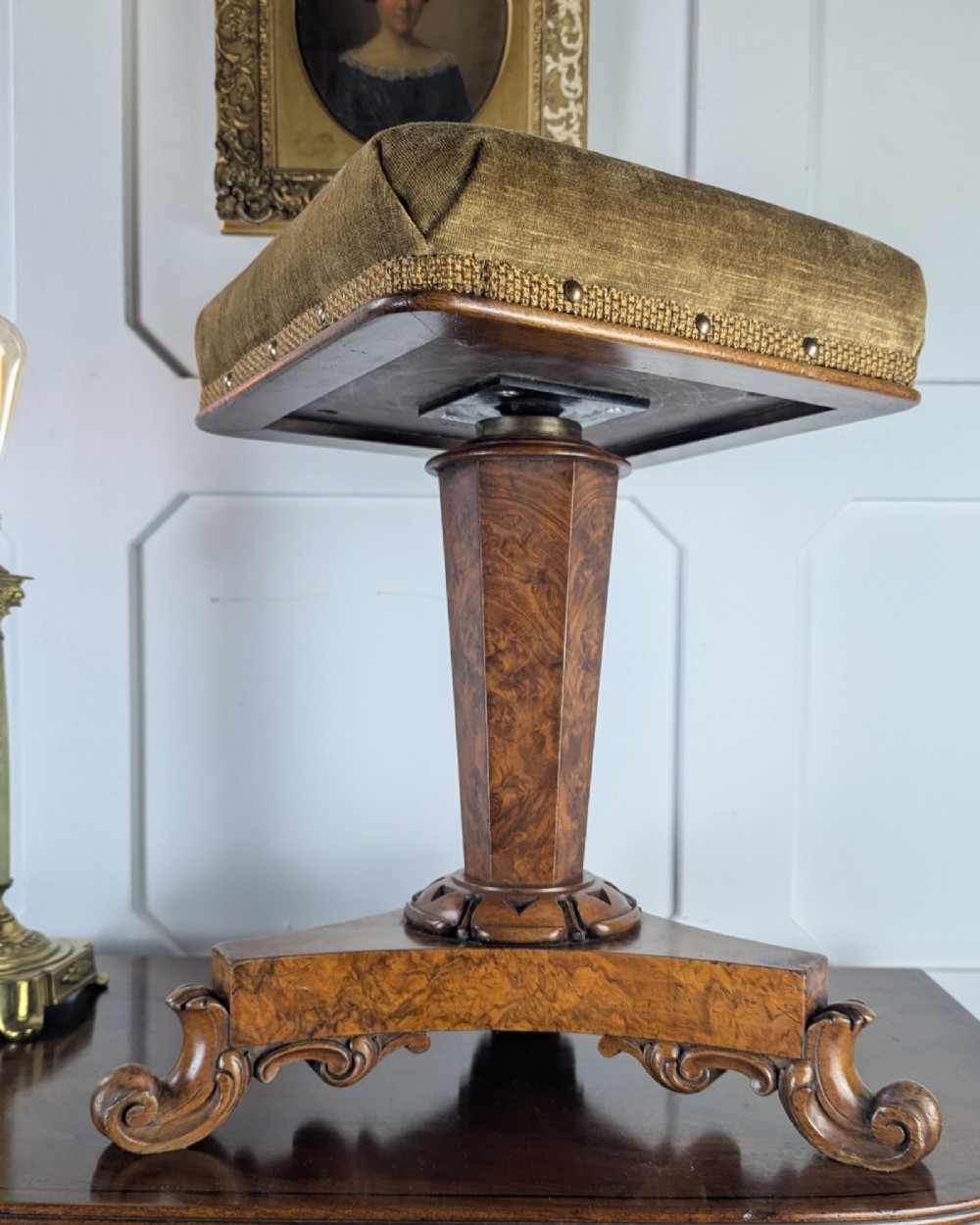 An antique William IV figured walnut revolving piano stool featuring an olive green upholstered square seat, a faceted column, and a carved triform base with scroll feet. View X