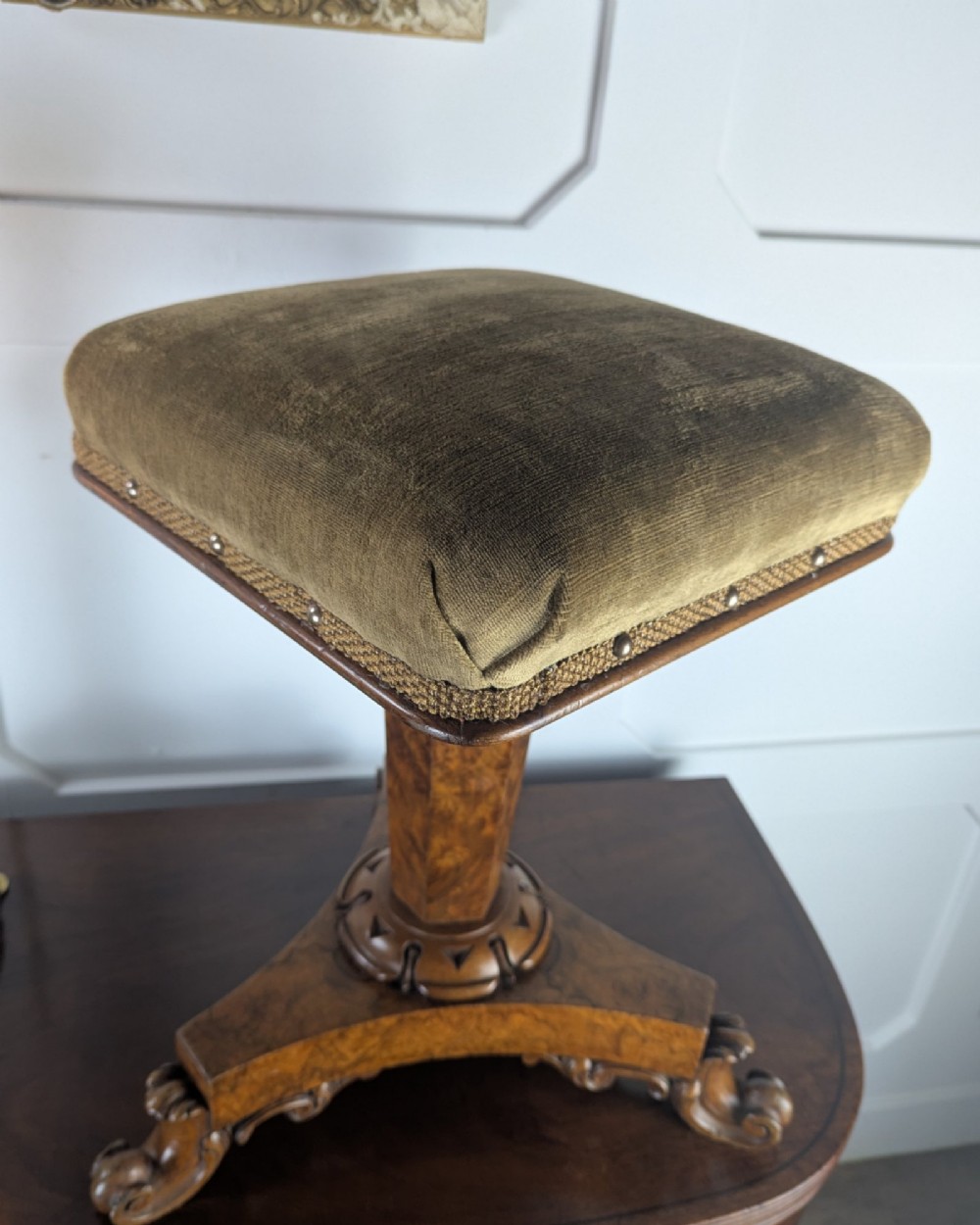 An antique William IV figured walnut revolving piano stool featuring an olive green upholstered square seat, a faceted column, and a carved triform base with scroll feet. View XII