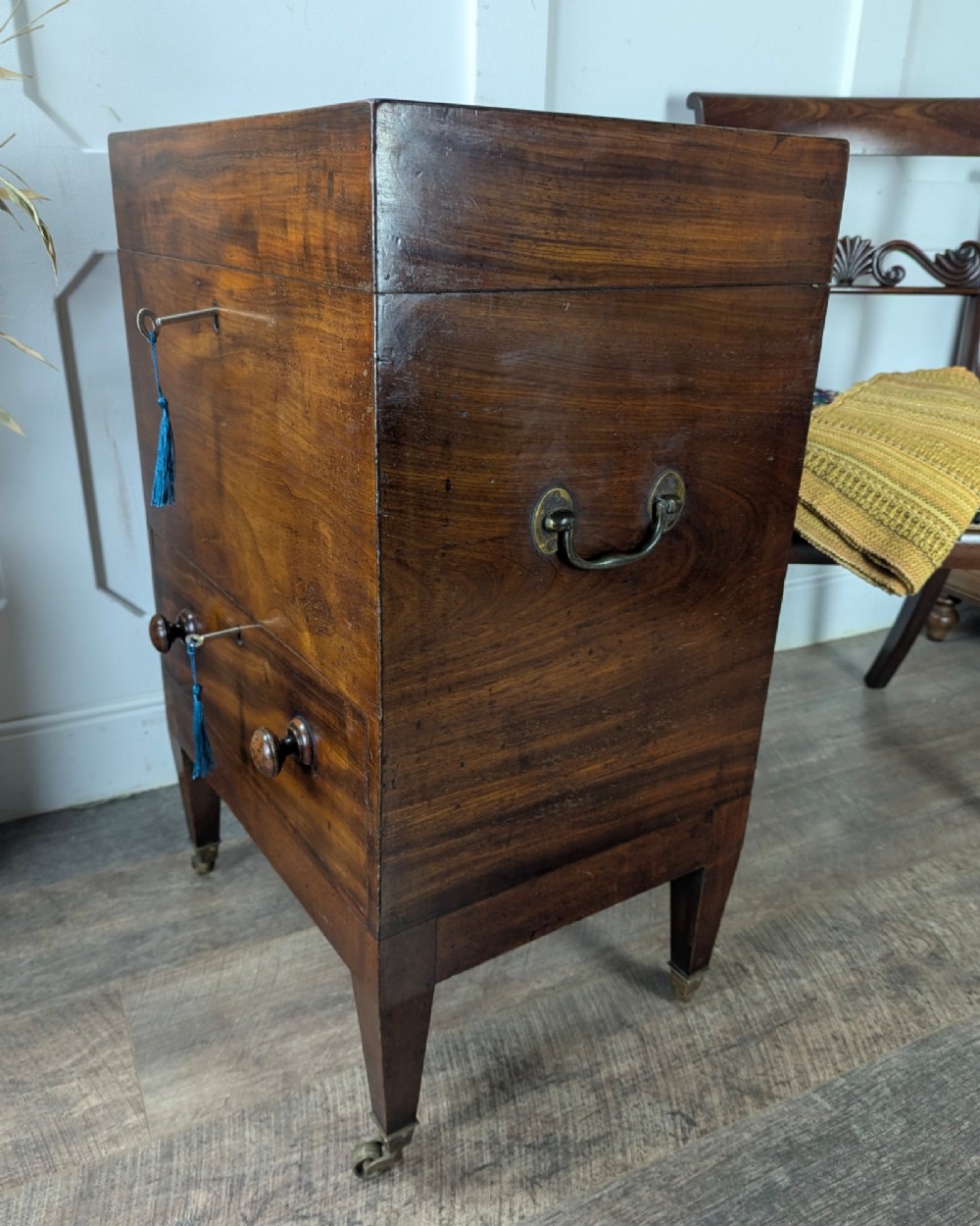 A Georgian dark mahogany cellarette featuring a hinged top lid, a lower drawer with wooden turned knobs, brass side handles, and square tapered legs on brass castors. View V