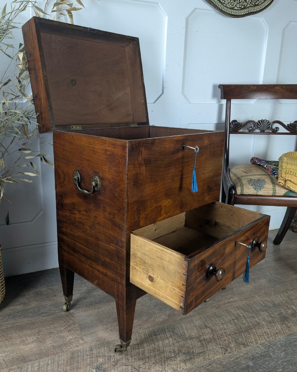 A Georgian dark mahogany cellarette featuring a hinged top lid, a lower drawer with wooden turned knobs, brass side handles, and square tapered legs on brass castors. View VI