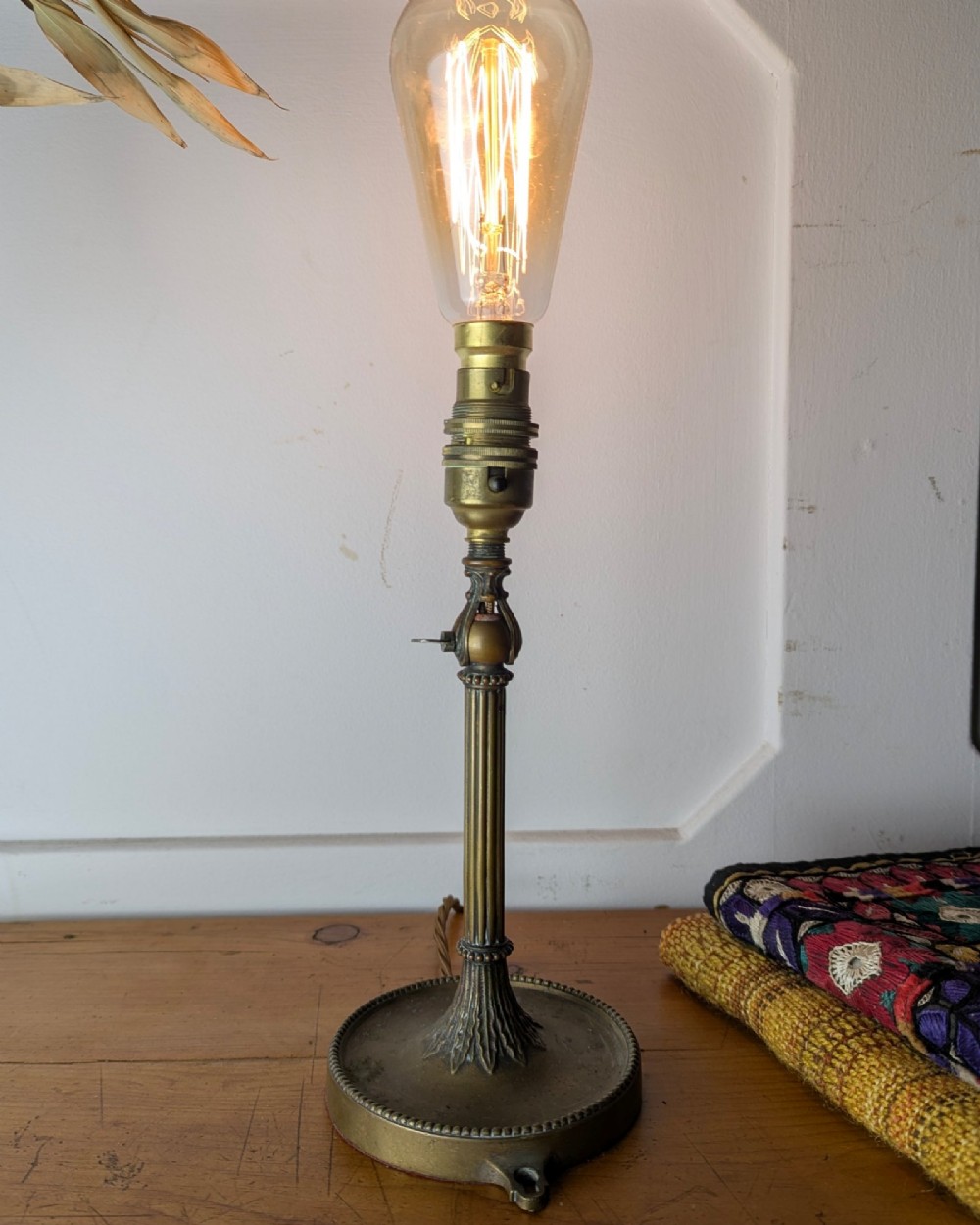 A slim early 20th-century aged brass lamp featuring a fluted stem, circular base, socket switch, and an exposed filament bulb. View II