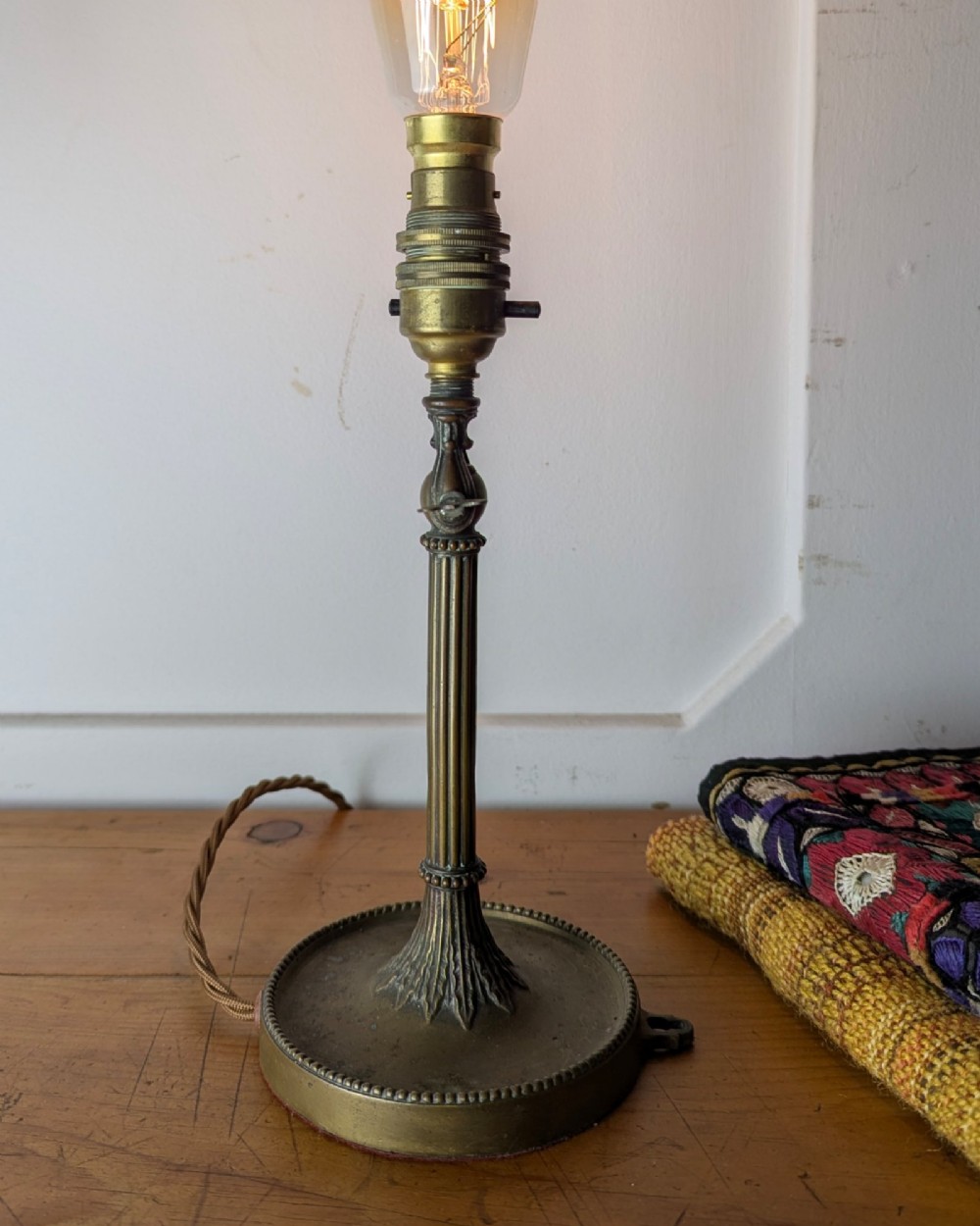 A slim early 20th-century aged brass lamp featuring a fluted stem, circular base, socket switch, and an exposed filament bulb. View IV