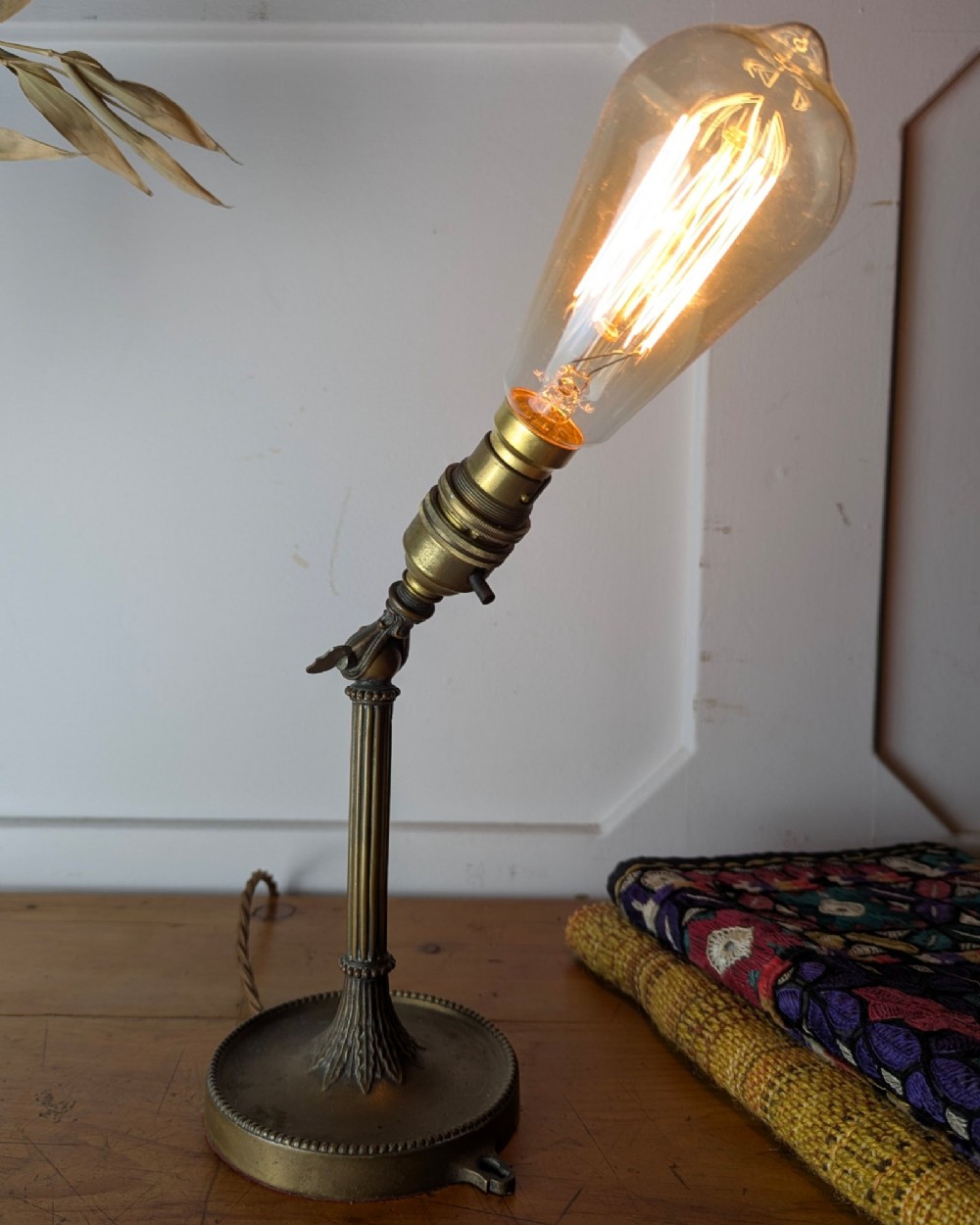 A slim early 20th-century aged brass lamp featuring a fluted stem, circular base, socket switch, and an exposed filament bulb. View V