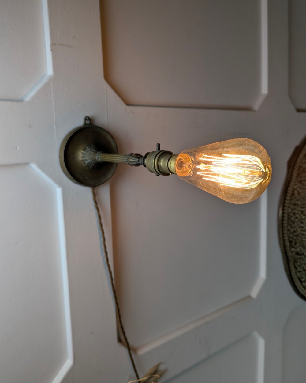 A slim early 20th-century aged brass lamp featuring a fluted stem, circular base, socket switch, and an exposed filament bulb. View VI