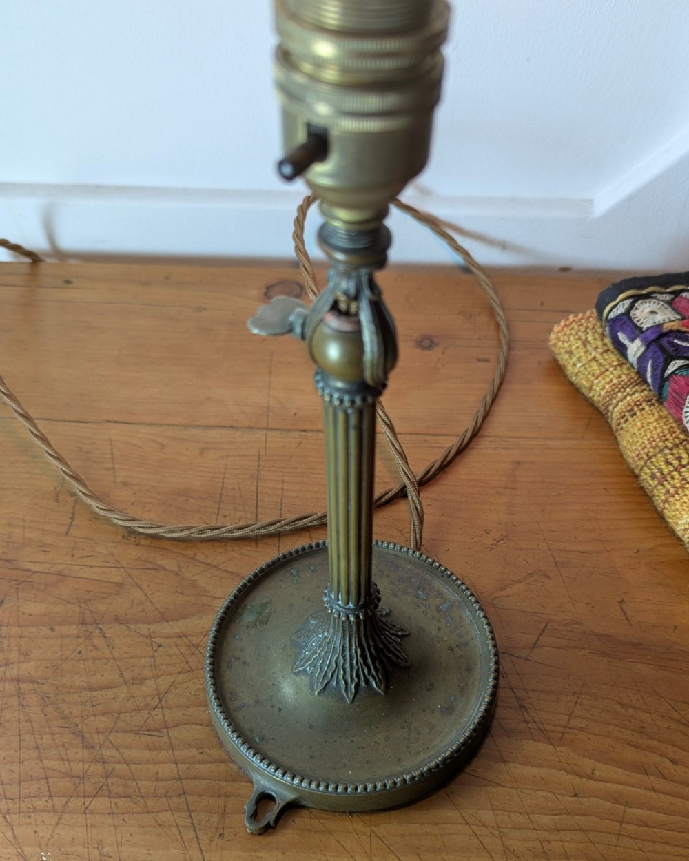 A slim early 20th-century aged brass lamp featuring a fluted stem, circular base, socket switch, and an exposed filament bulb. View VII