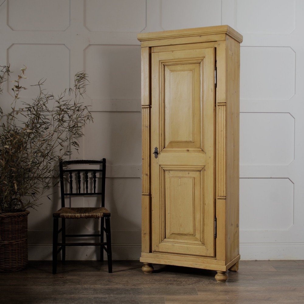 A tall single door stripped pine armoire featuring fielded panels, fluted side pilasters, and turned bun feet.