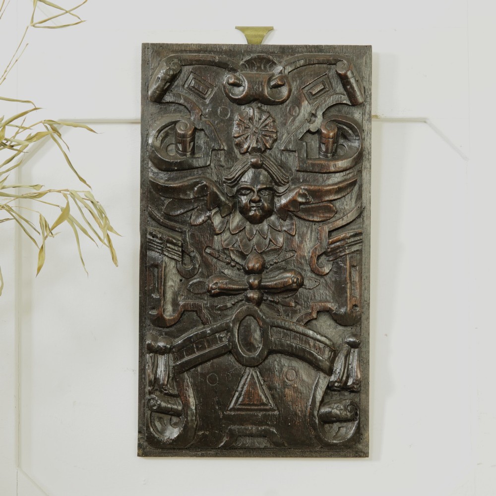 A rectangular dark oak timber panel featuring a high-relief hand carving of a winged cherub head surrounded by intricate scrollwork and geometric strapwork.