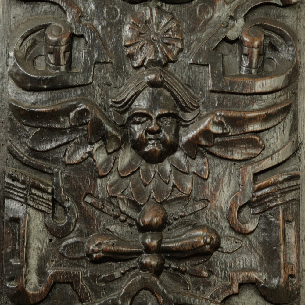 A rectangular dark oak timber panel featuring a high-relief hand carving of a winged cherub head surrounded by intricate scrollwork and geometric strapwork. View V