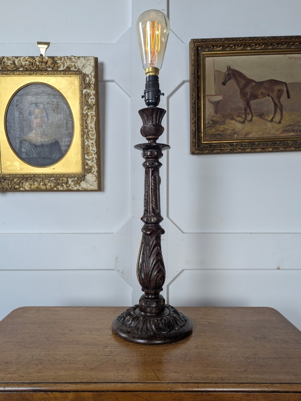 A late Victorian carved dark oak candlestick table lamp base featuring detailed acanthus leaf carvings and an exposed filament bulb.