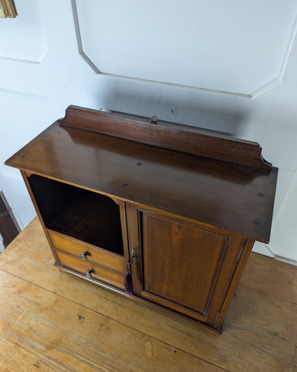 A late nineteenth-century warm brown wooden smokers cabinet featuring an open shelf over two drawers on the left, a single inlaid door on the right, and brass hardware. View VI