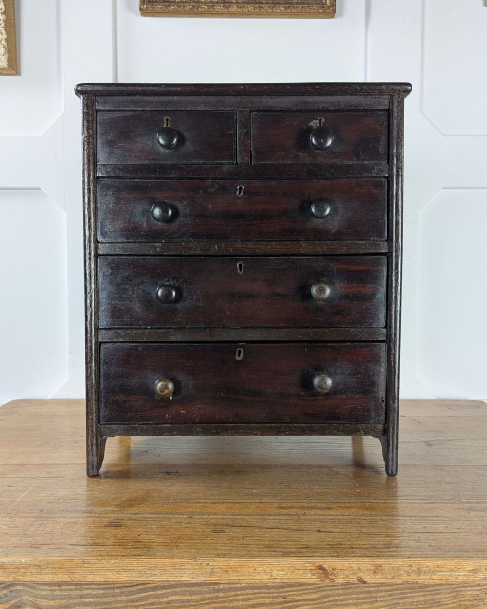 A 19th-century miniature wooden chest of drawers with a dark stained finish, two small top drawers over three long drawers, turned knobs, and brass escutcheons. View VI