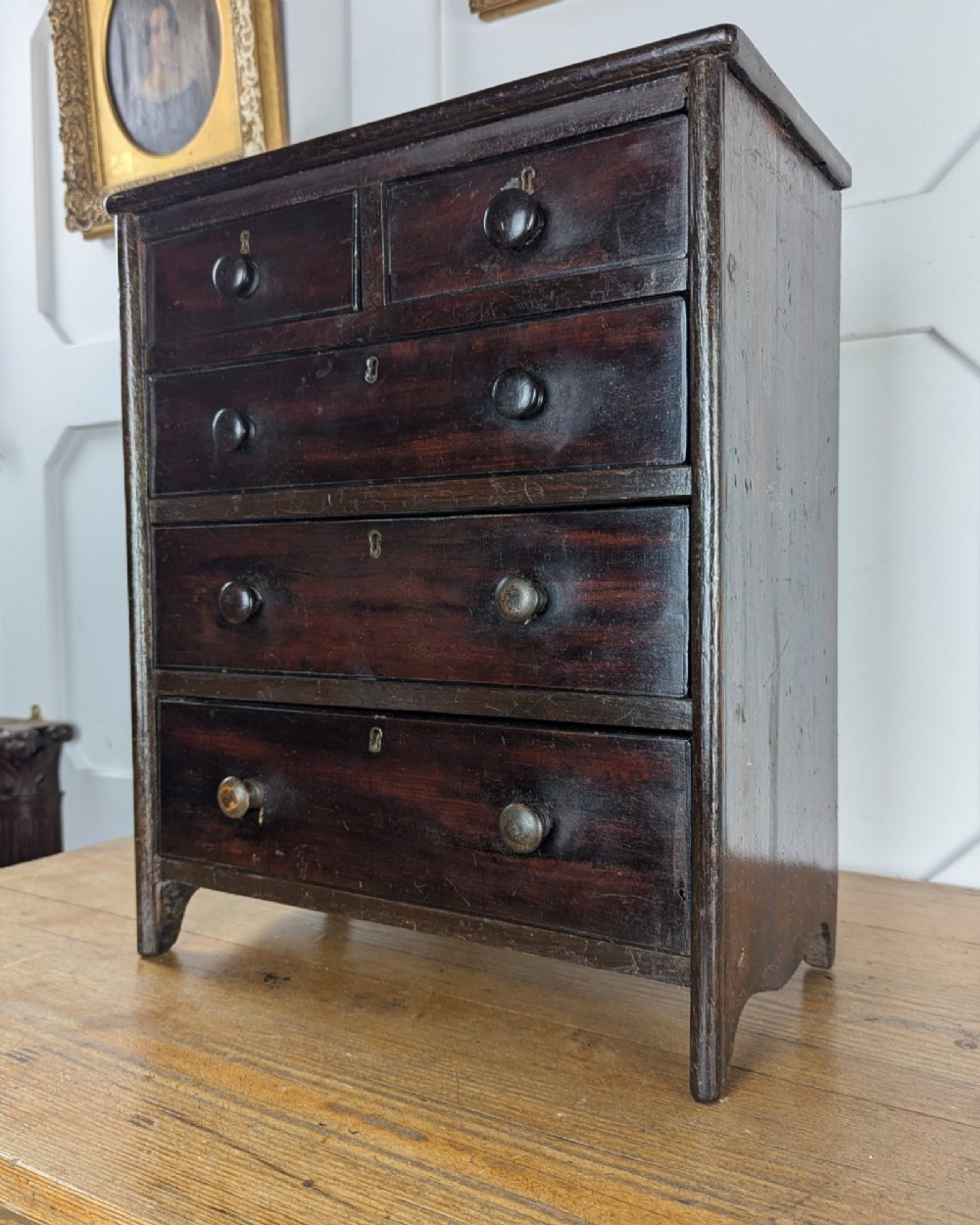 A 19th-century miniature wooden chest of drawers with a dark stained finish, two small top drawers over three long drawers, turned knobs, and brass escutcheons. View VII