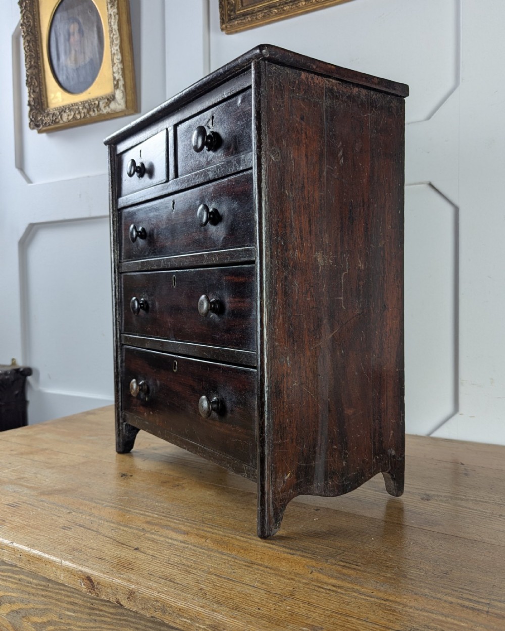 A 19th-century miniature wooden chest of drawers with a dark stained finish, two small top drawers over three long drawers, turned knobs, and brass escutcheons. View VIII