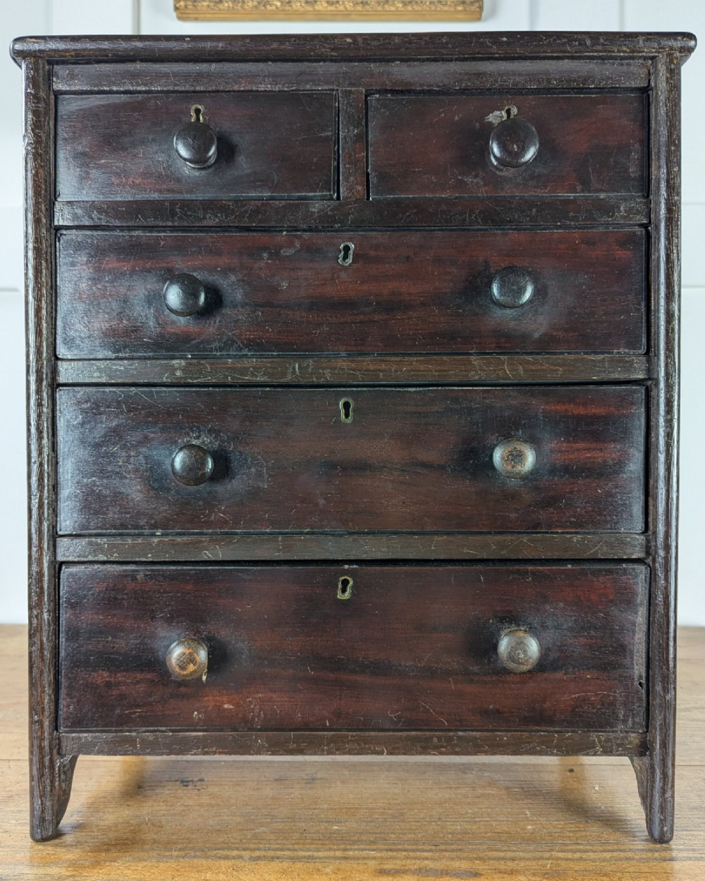 A 19th-century miniature wooden chest of drawers with a dark stained finish, two small top drawers over three long drawers, turned knobs, and brass escutcheons. View IX