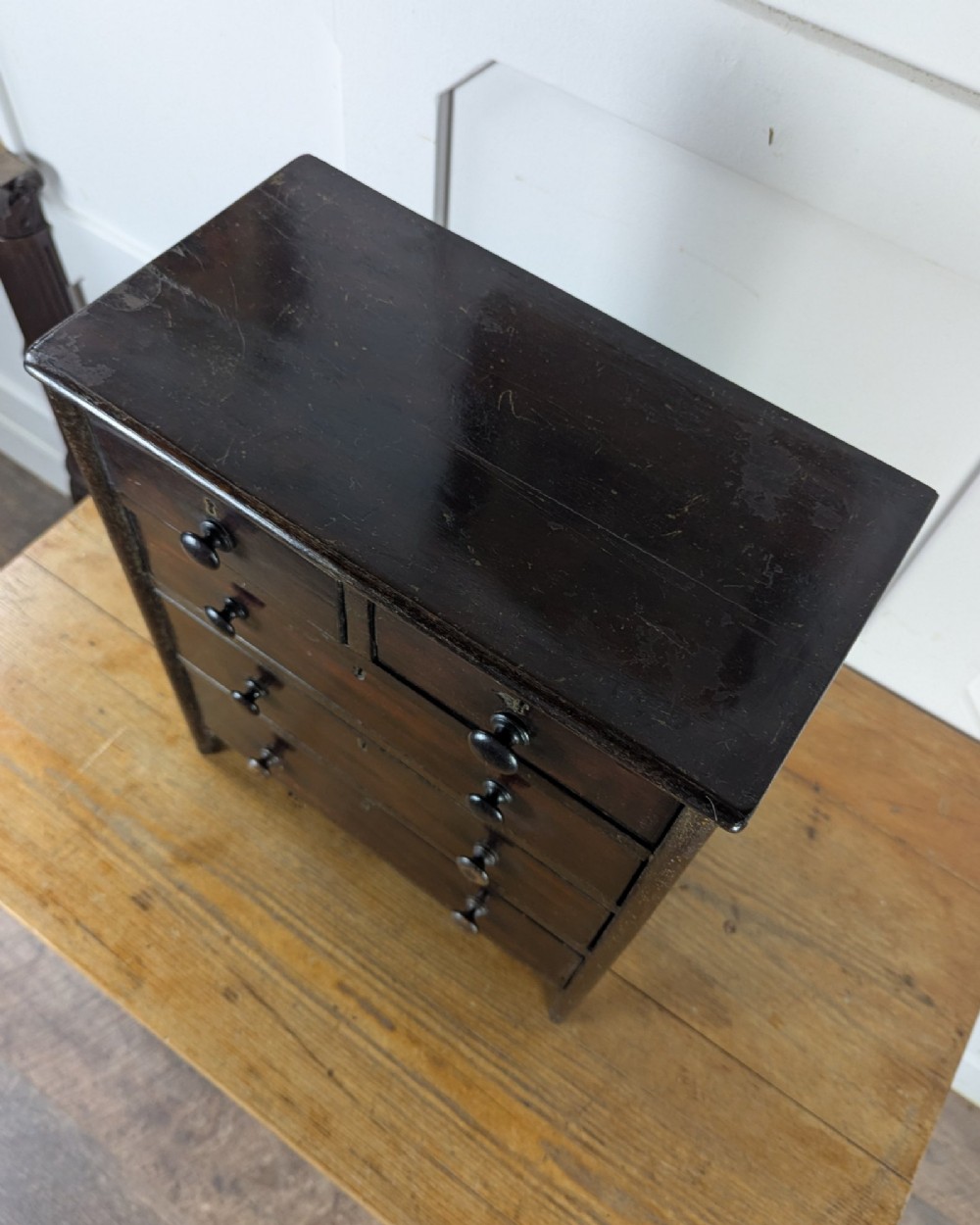 A 19th-century miniature wooden chest of drawers with a dark stained finish, two small top drawers over three long drawers, turned knobs, and brass escutcheons. View XII