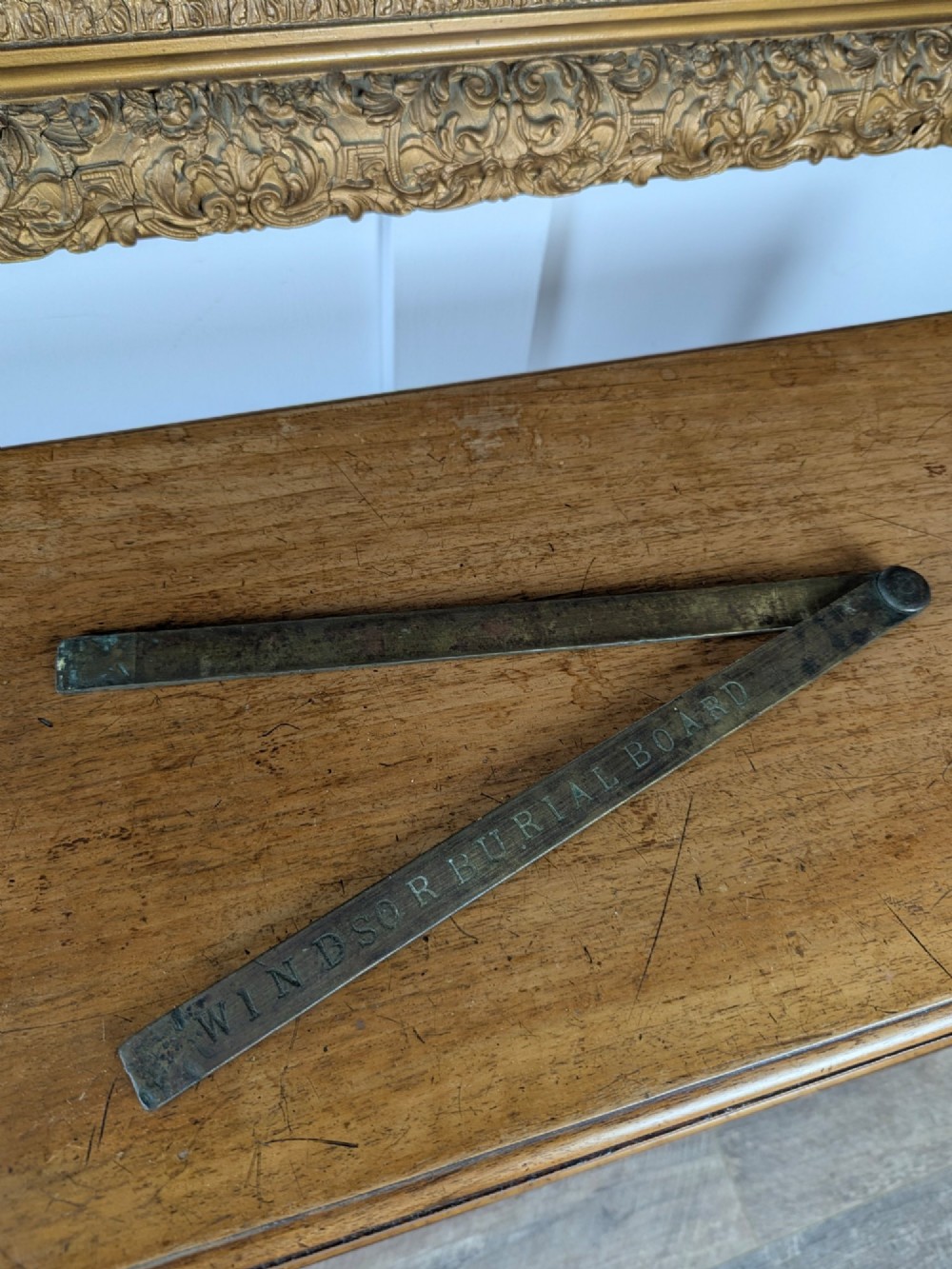 A solid brass folding ruler featuring stamped block text reading WINDSOR BURIAL BOARD resting on a wooden surface. View II