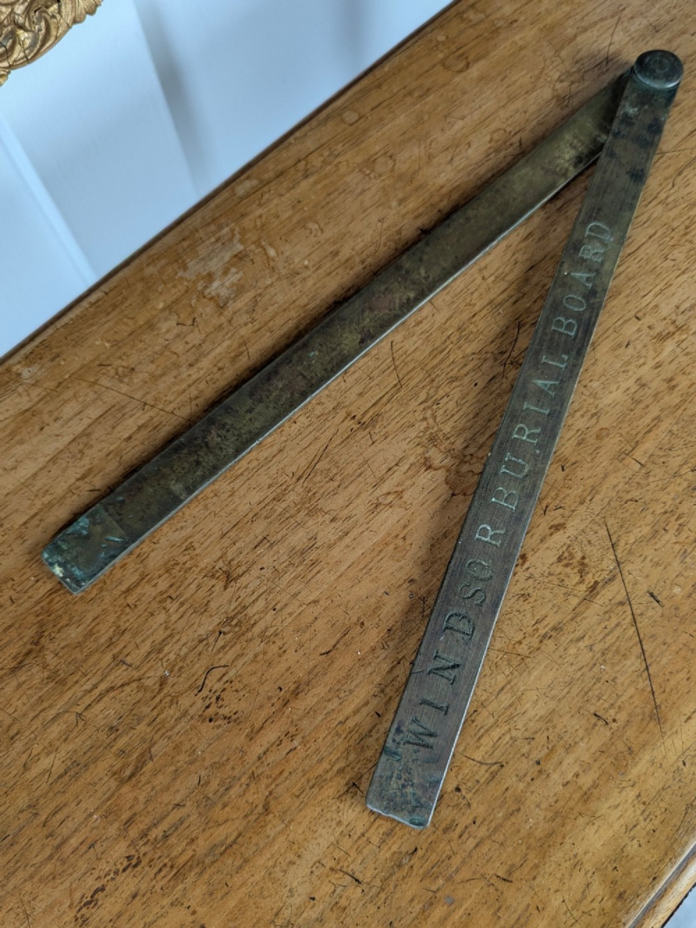 A solid brass folding ruler featuring stamped block text reading WINDSOR BURIAL BOARD resting on a wooden surface. View V