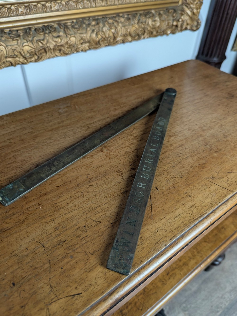 A solid brass folding ruler featuring stamped block text reading WINDSOR BURIAL BOARD resting on a wooden surface. View VI