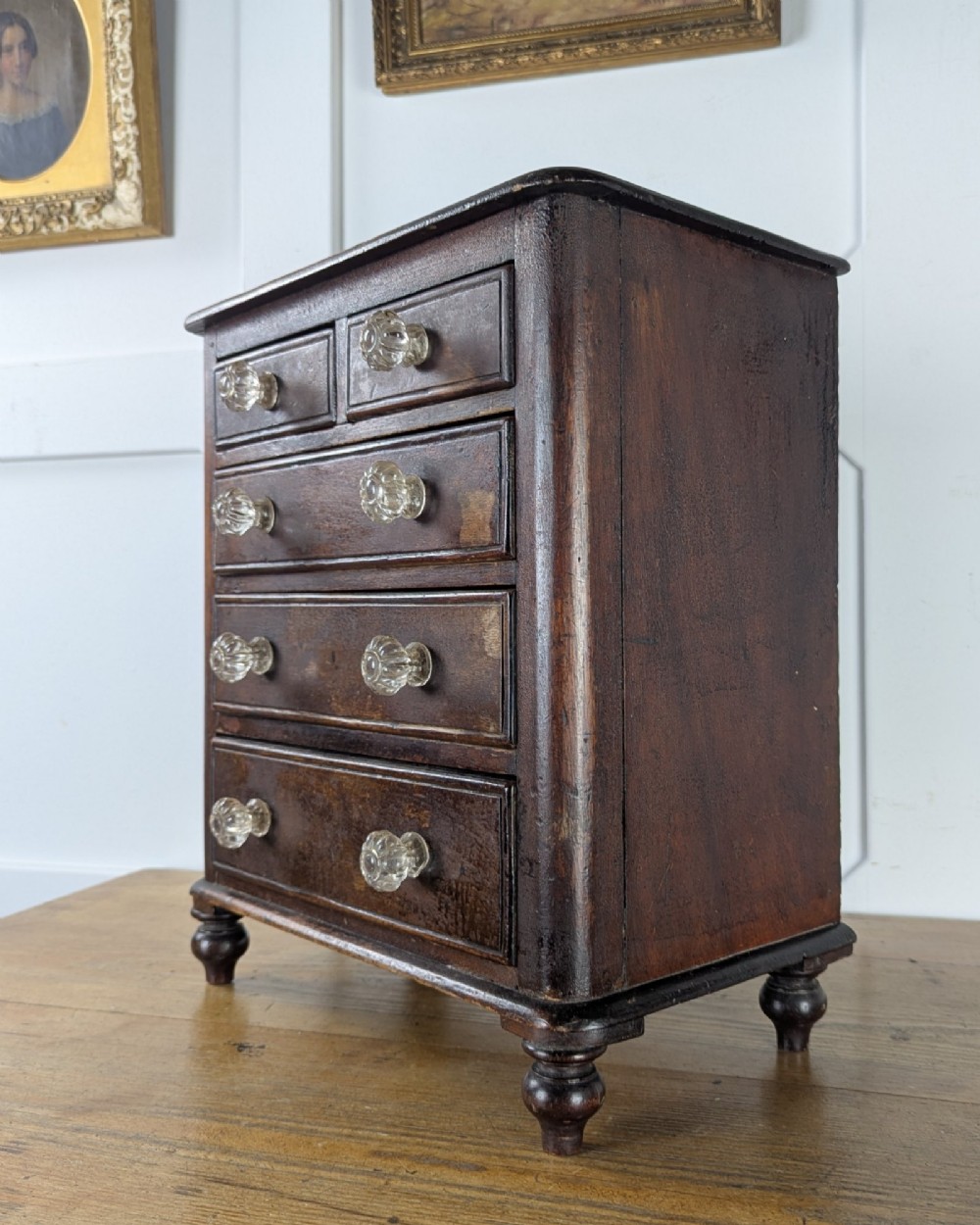 A dark brown Victorian miniature wooden chest of drawers with glass knobs and small turned feet. View II