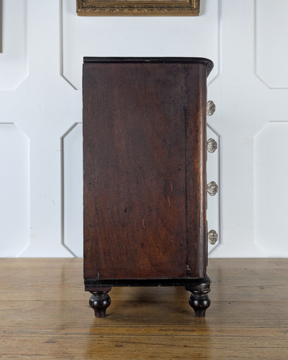 A dark brown Victorian miniature wooden chest of drawers with glass knobs and small turned feet. View V