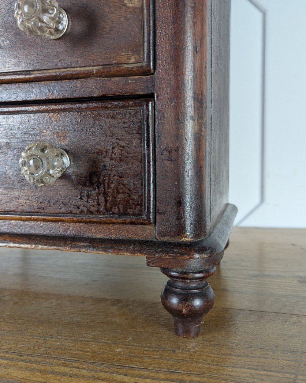A dark brown Victorian miniature wooden chest of drawers with glass knobs and small turned feet. View VI