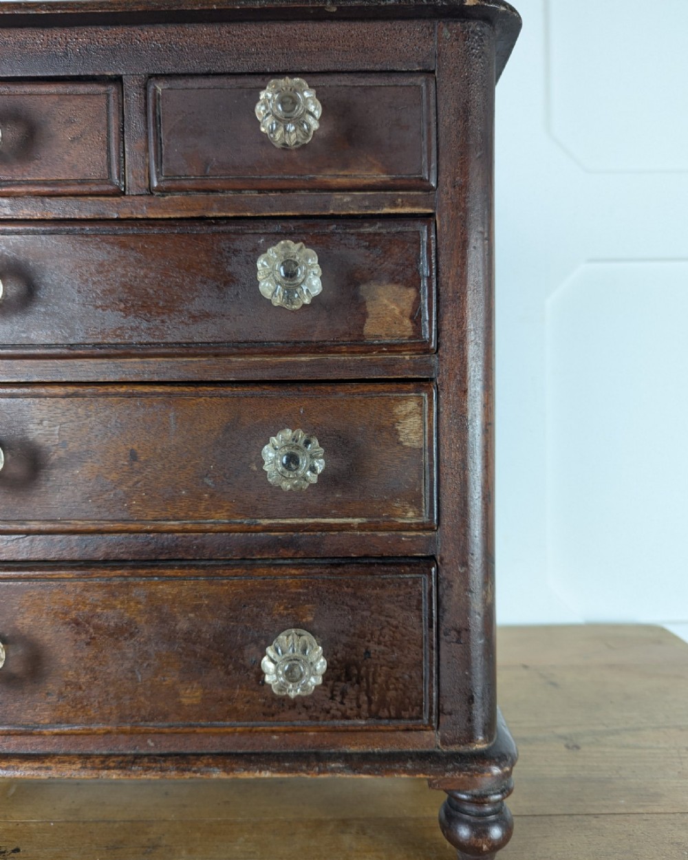 A dark brown Victorian miniature wooden chest of drawers with glass knobs and small turned feet. View VII