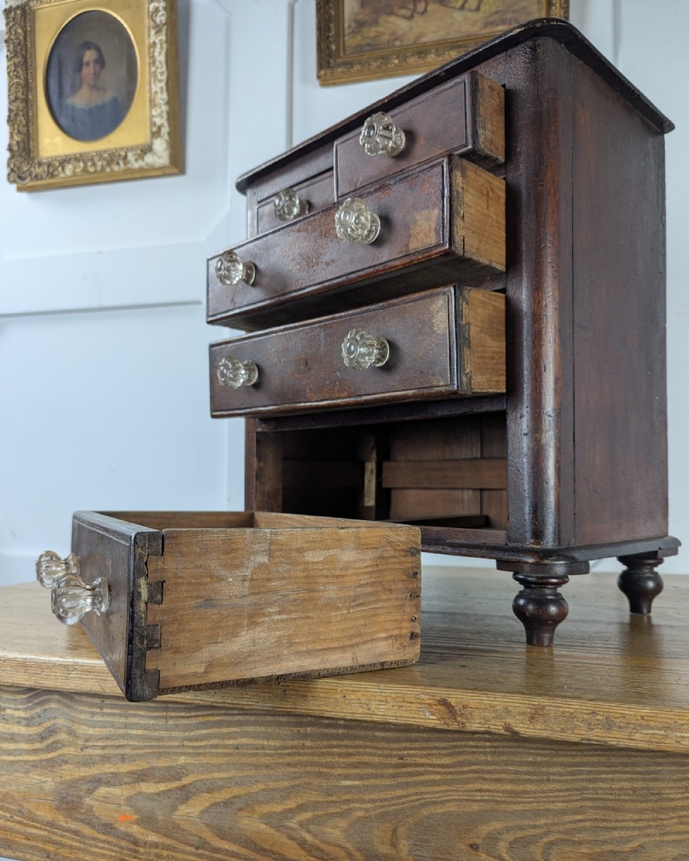 A dark brown Victorian miniature wooden chest of drawers with glass knobs and small turned feet. View X