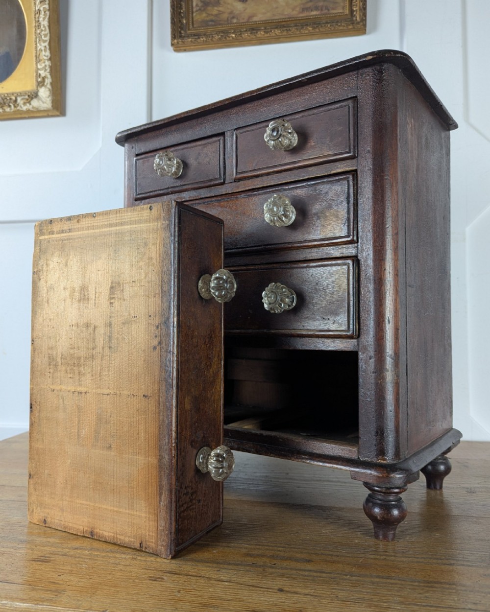 A dark brown Victorian miniature wooden chest of drawers with glass knobs and small turned feet. View XI